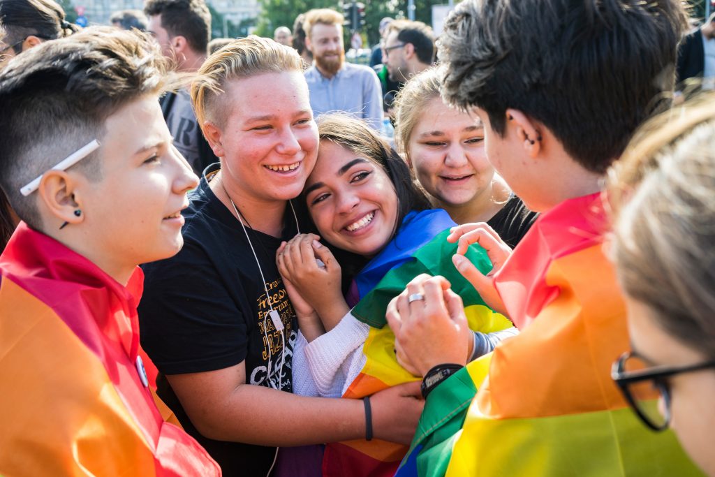Older children smile and hug while wearing the Pride flag. 
