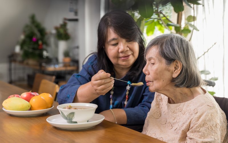 A woman feeds a senior citzen a bowl of soup while sitting at a dining room table.
