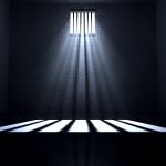 An old jail cell interior with barred up window with light rays penetrating through it reflecting on the floor.