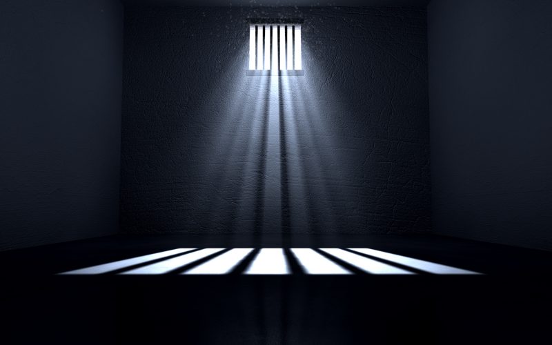 An old jail cell interior with barred up window with light rays penetrating through it reflecting on the floor.