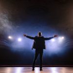 A person stands on a stage with his arms open wide soaking in adulation from the crowd.