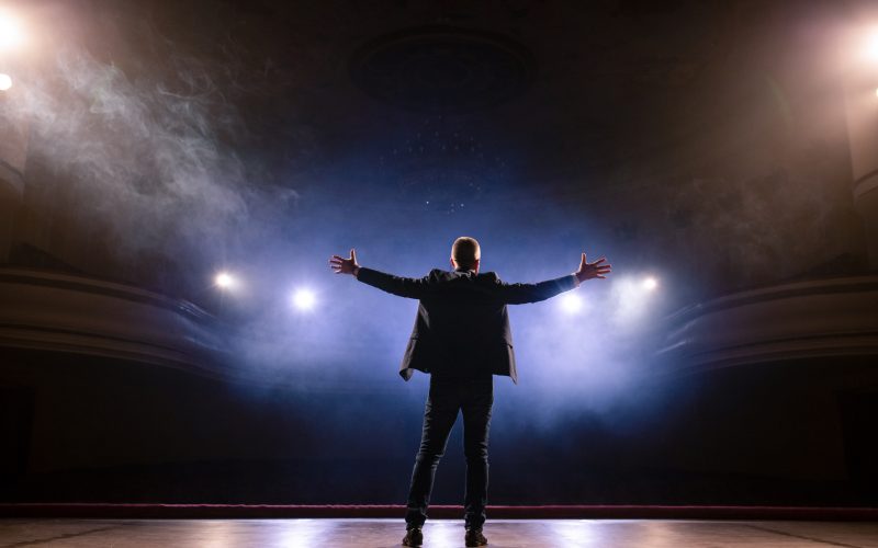 A person stands on a stage with his arms open wide soaking in adulation from the crowd.